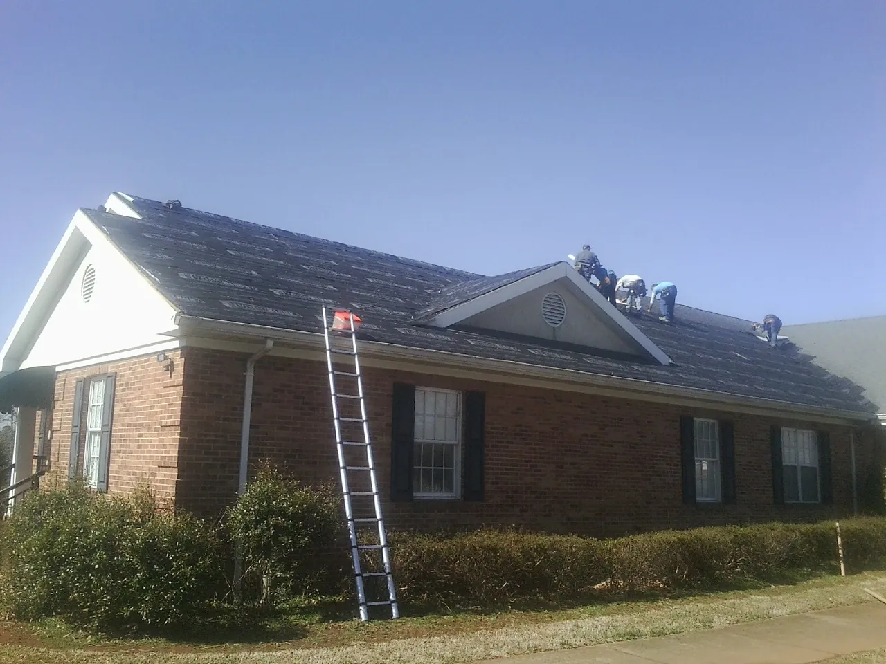 Crew tearing shingles off a brick ranch roof with a ladder set — Emergency Roof Repair work by our crew in West Ossipee