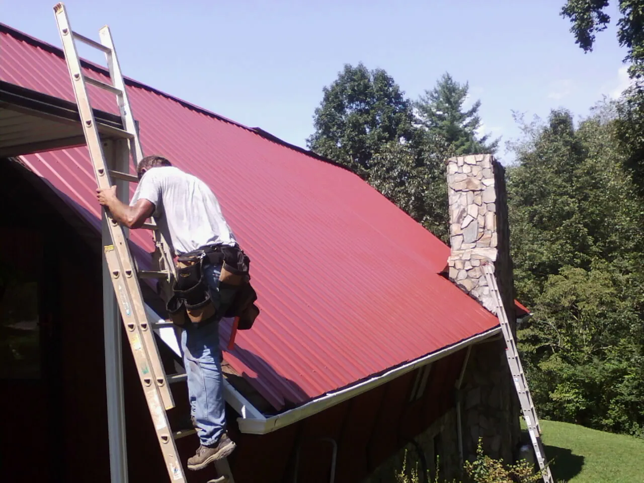 Roofer on a ladder beside a red metal roof and stone chimney — Soffit & Fascia Repair by Roofing Brothers in West Ossipee